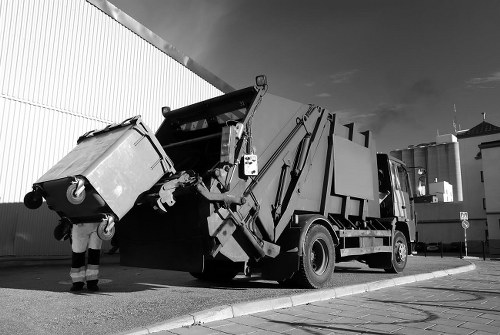 A large industrial waste collection truck is parked on a paved street outside a warehouse or industrial building, with a high metal wall to the left and clear sky above. The truck features a hydraulic lifting mechanism at the rear, which is actively engaged in emptying a large, rectangular, wheeled waste bin made of durable plastic or metal, with a textured surface and a lid that is partially open. The waste bin is suspended at an angle as it is being emptied into the truck's hopper. Nearby, a person wearing work trousers and safety boots stands close to the bin, observing the process. The scene captures the essence of commercial waste collection services, with the truck designed for efficient rubbish removal and disposal in an urban or industrial setting, supporting companies like Garden Clearance Sudbury in managing rubbish removal tasks.