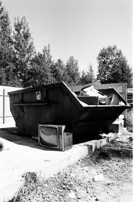 A large, open-top metal skip container with a dark, weathered surface and visible rust spots, positioned on a concrete paving slab in an outdoor area. The container is filled with miscellaneous bulky 